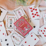 A set of Christmas-themed playing cards with festive illustrations and a red patterned back, arranged on a soft beige fabric background with blurred holiday lights and greenery.