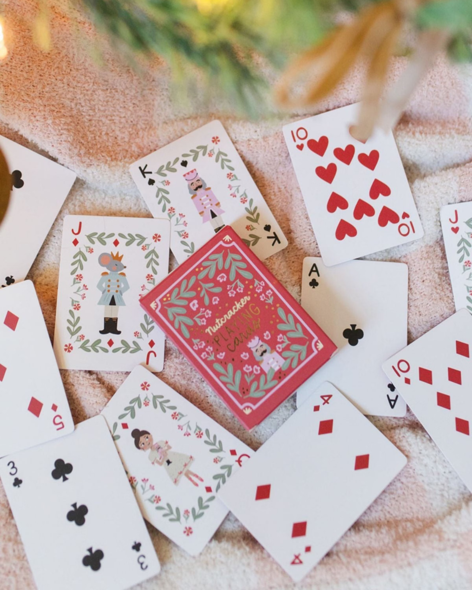 A set of Christmas-themed playing cards with festive illustrations and a red patterned back, arranged on a soft beige fabric background with blurred holiday lights and greenery.