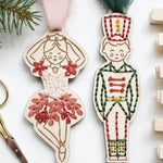 Two wooden Christmas ornaments shaped like a ballerina and a nutcracker, decorated with colorful embroidered details, displayed on a white surface with scissors, pine branches, and wooden clothespins.