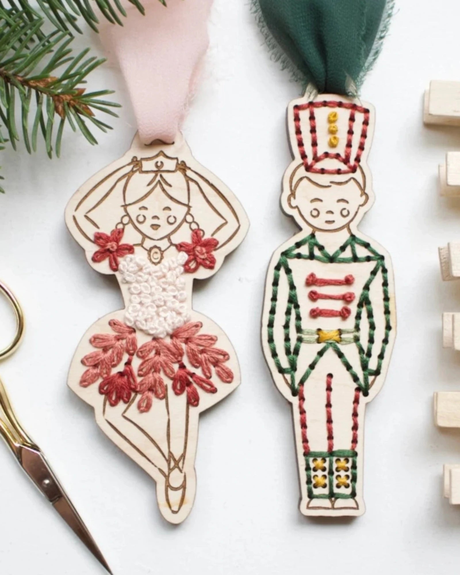 Two wooden Christmas ornaments shaped like a ballerina and a nutcracker, decorated with colorful embroidered details, displayed on a white surface with scissors, pine branches, and wooden clothespins.
