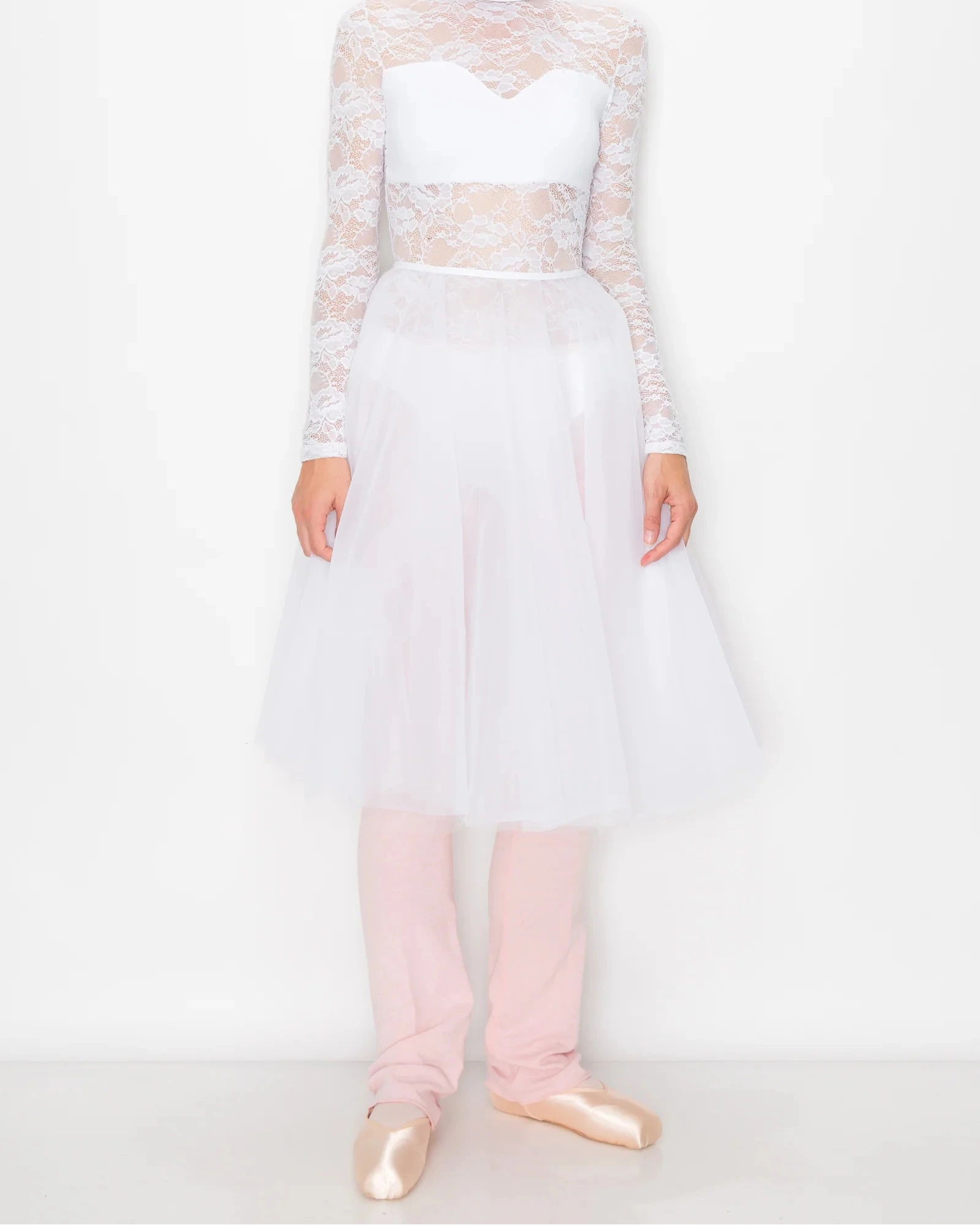 A model wearing a white lace and tulle ballet outfit with long sleeves, a sweetheart bodice, and a full tutu skirt, paired with light pink leggings and metallic pink ballet slippers, against a plain white studio background.