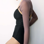 A woman wearing a black leotard with a long-sleeve, semi-sheer mauve-brown rash guard layered over her shoulders, posing against a plain light gray wall.