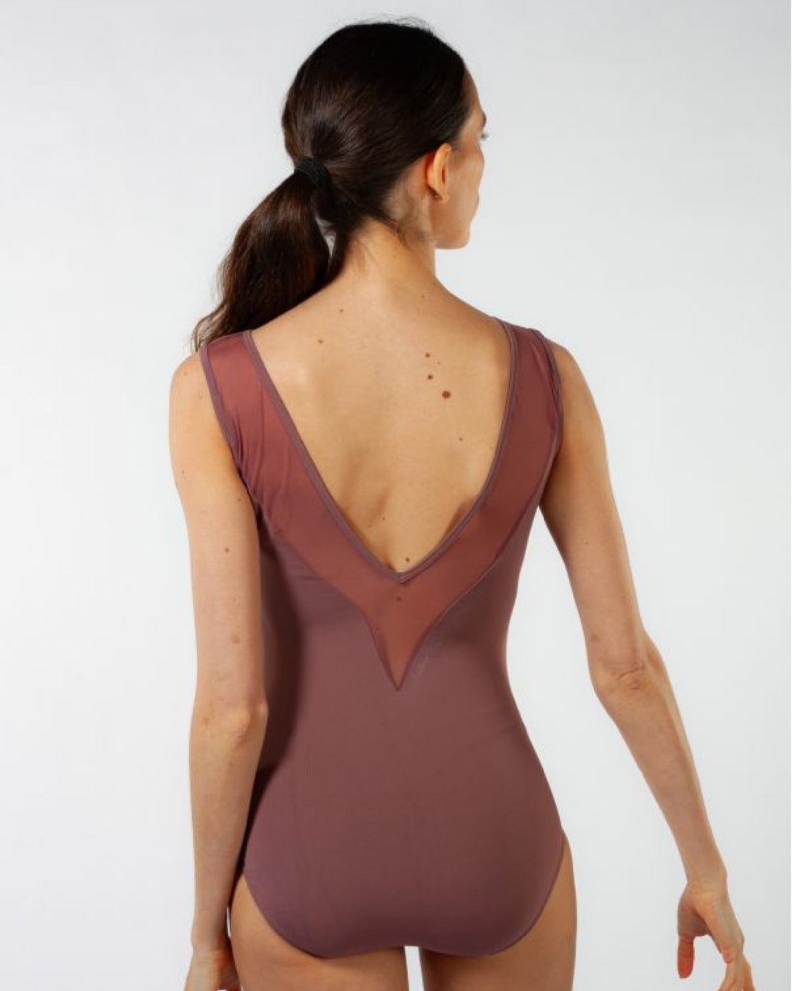 A woman shown from the back wearing a sleeveless leotard in a muted dusty rose or mauve color with sheer mesh paneling at the back, photographed against a clean light gray studio background.