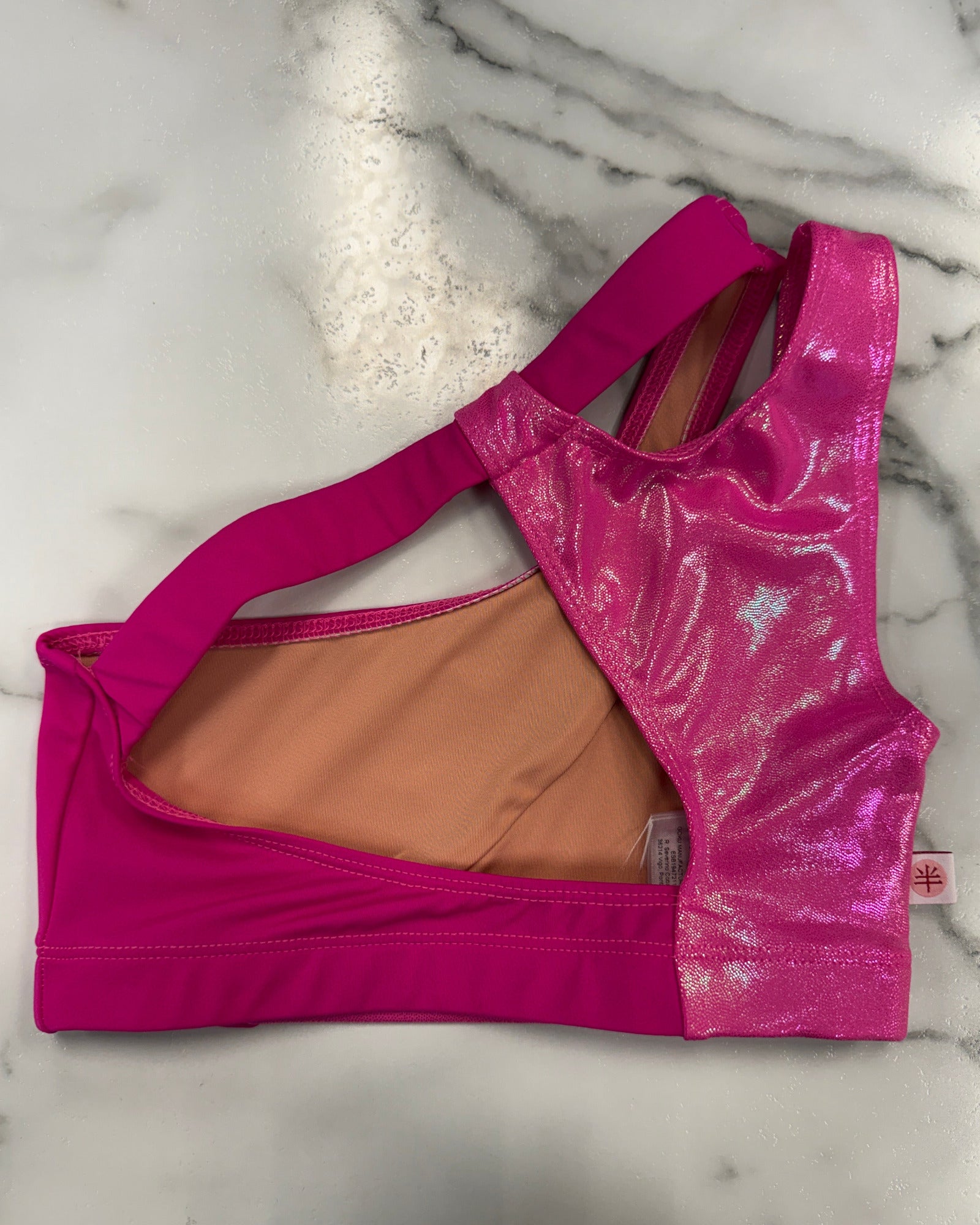 A bright pink dance bra with a shiny holographic mesh back panel and a peach-colored lining, laid flat on a white marble surface.