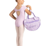 A young girl dancer in a light purple leotard and tights stands in profile holding a lavender and white sequined dance bag with the word 'dance' written on it, against a solid white background.