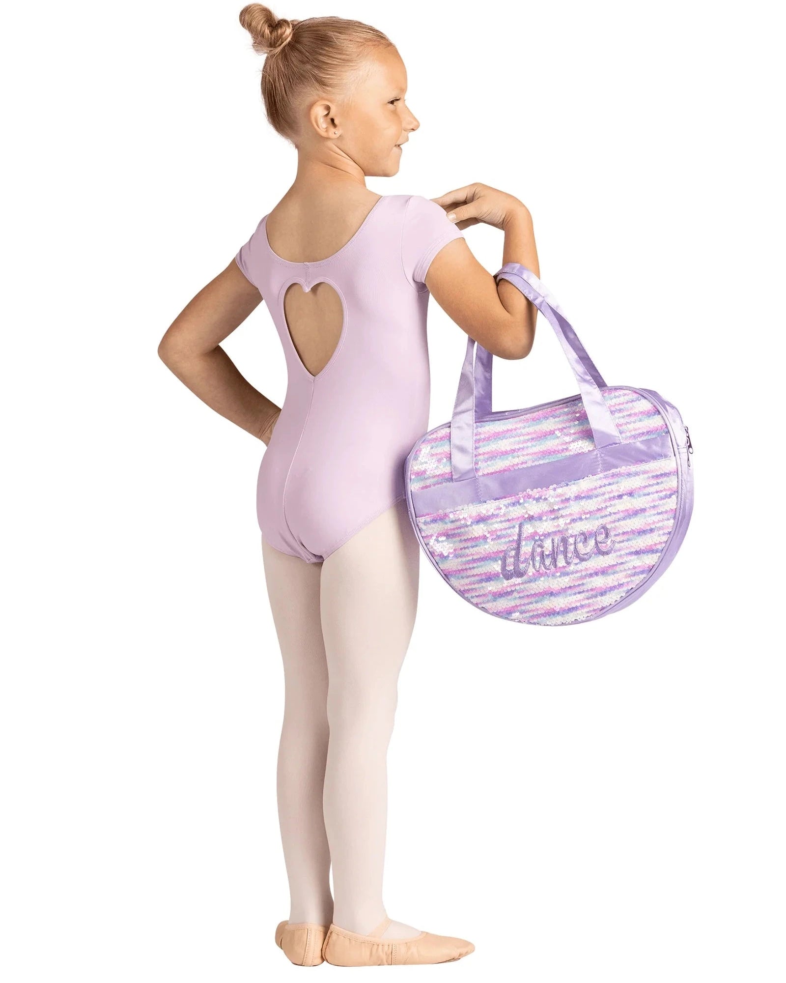 A young girl dancer in a light purple leotard and tights stands in profile holding a lavender and white sequined dance bag with the word 'dance' written on it, against a solid white background.
