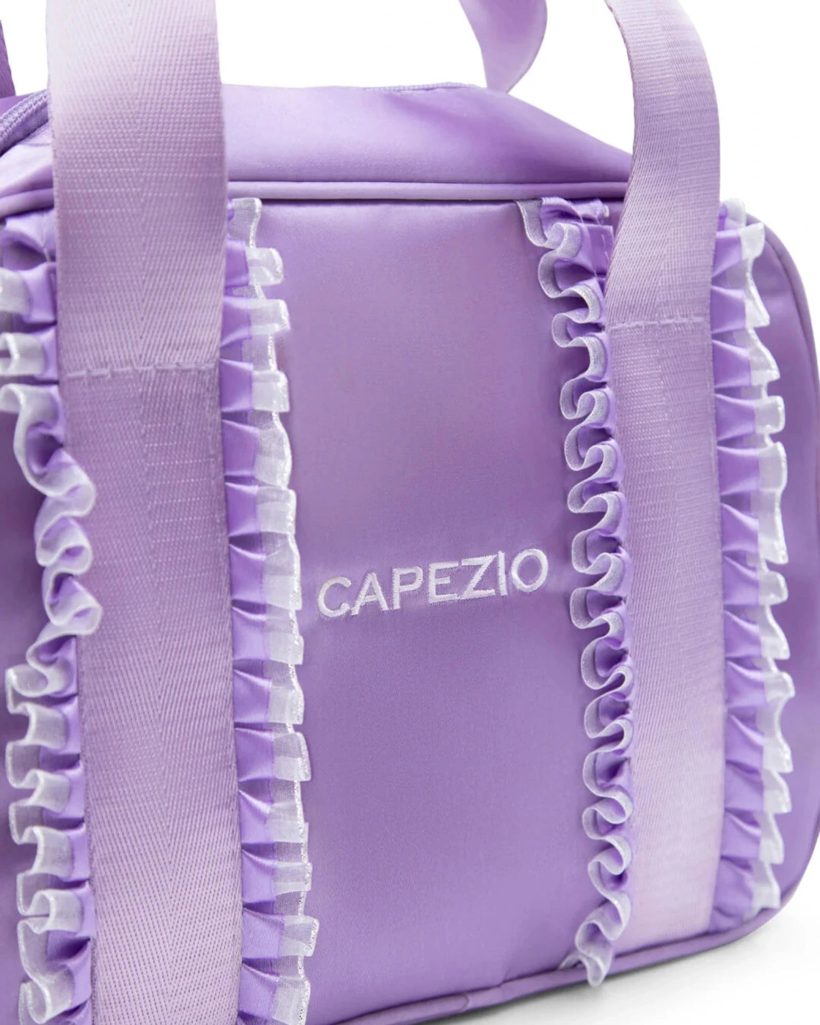 A close-up of a lavender purple dance bag with ruffled trim and wide shoulder straps, featuring the brand name 'CAPEZIO' printed in white on the front.