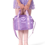 A child wearing a lavender tutu skirt and ballet slippers holds a shiny lavender dance bag with a shoulder strap. The bag has decorative ruffle trim and a small white logo that reads 'DANZO'.