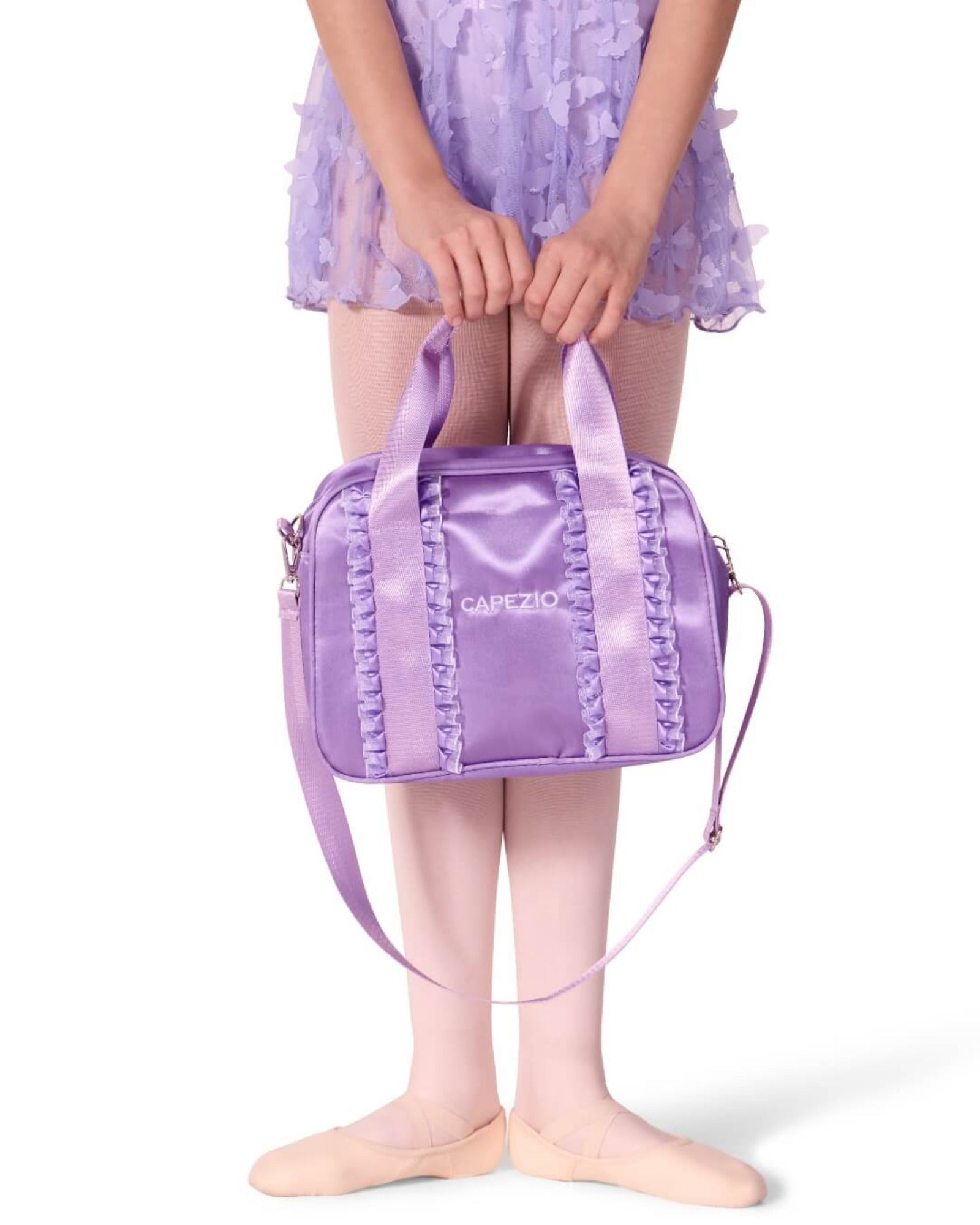 A child wearing a lavender tutu skirt and ballet slippers holds a shiny lavender dance bag with a shoulder strap. The bag has decorative ruffle trim and a small white logo that reads 'DANZO'.