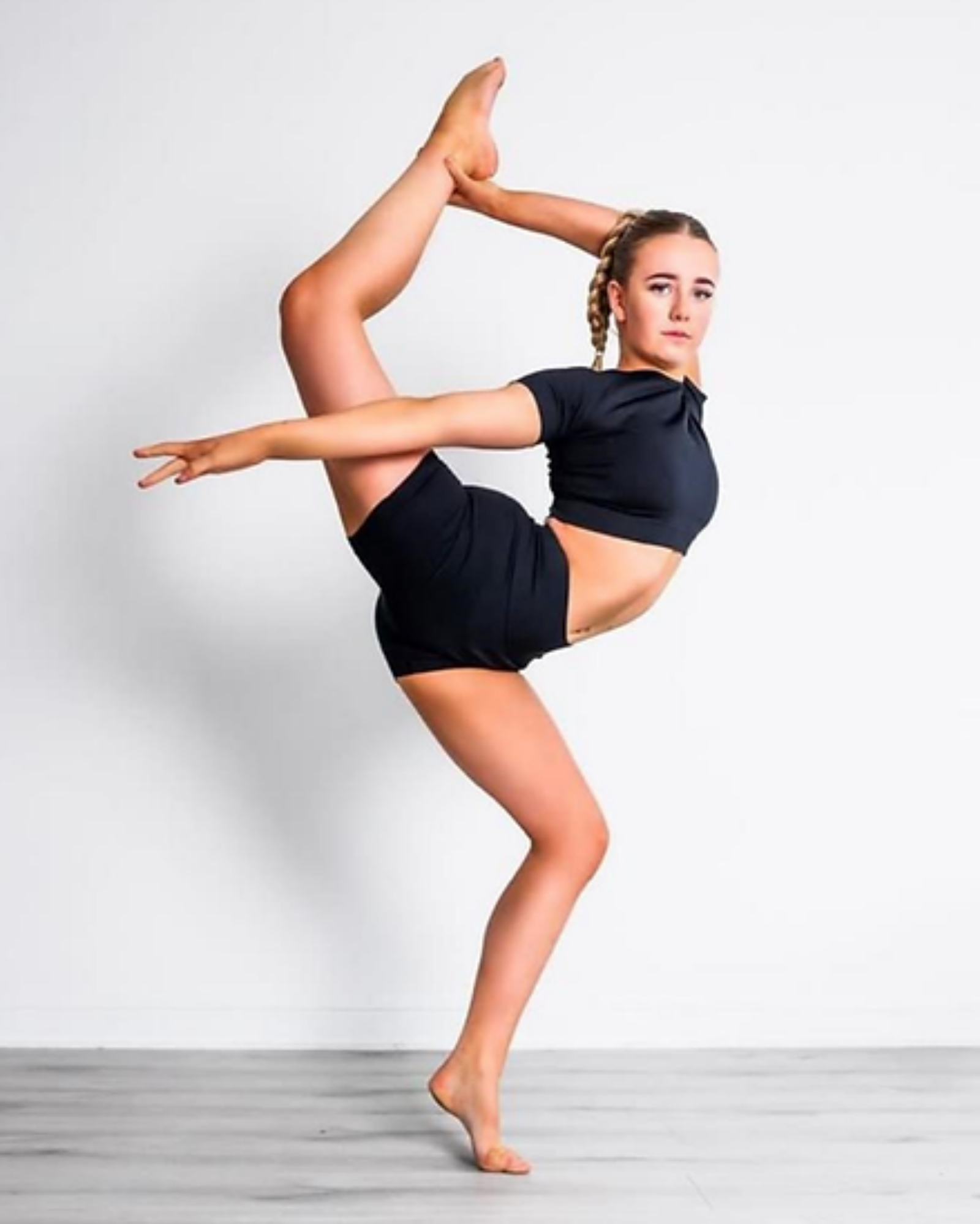A young female dancer in a black crop top and matching shorts performs a high leg lift against a plain white studio background.