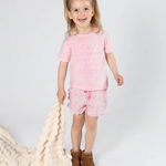 A young girl with blonde hair in a ponytail stands against a light gray studio background, wearing a light pink pajama shirt and matching pink patterned shorts. She is holding a chunky cream knit blanket and wearing brown boots.