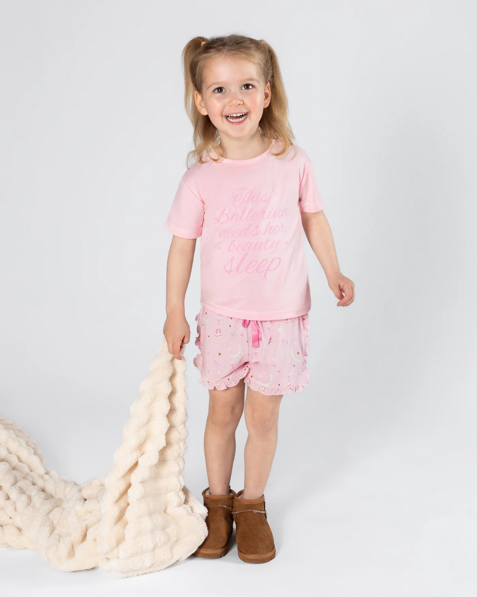 A young girl with blonde hair in a ponytail stands against a light gray studio background, wearing a light pink pajama shirt and matching pink patterned shorts. She is holding a chunky cream knit blanket and wearing brown boots.