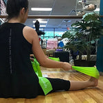 A woman sits on a wooden floor in a dance studio, using a bright green leg stretch band to stretch her leg. She is wearing a black tank top and black leggings.
