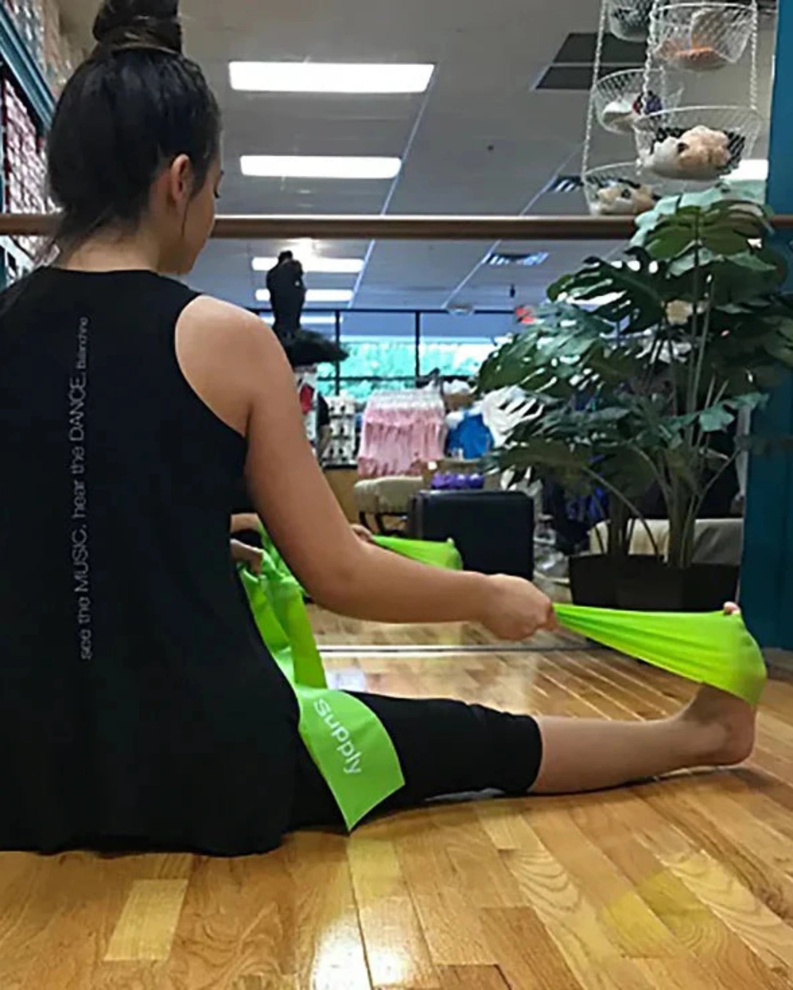 A woman sits on a wooden floor in a dance studio, using a bright green leg stretch band to stretch her leg. She is wearing a black tank top and black leggings.