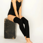 Woman sitting on a black block wearing a black sleeveless top and leggings against a white background