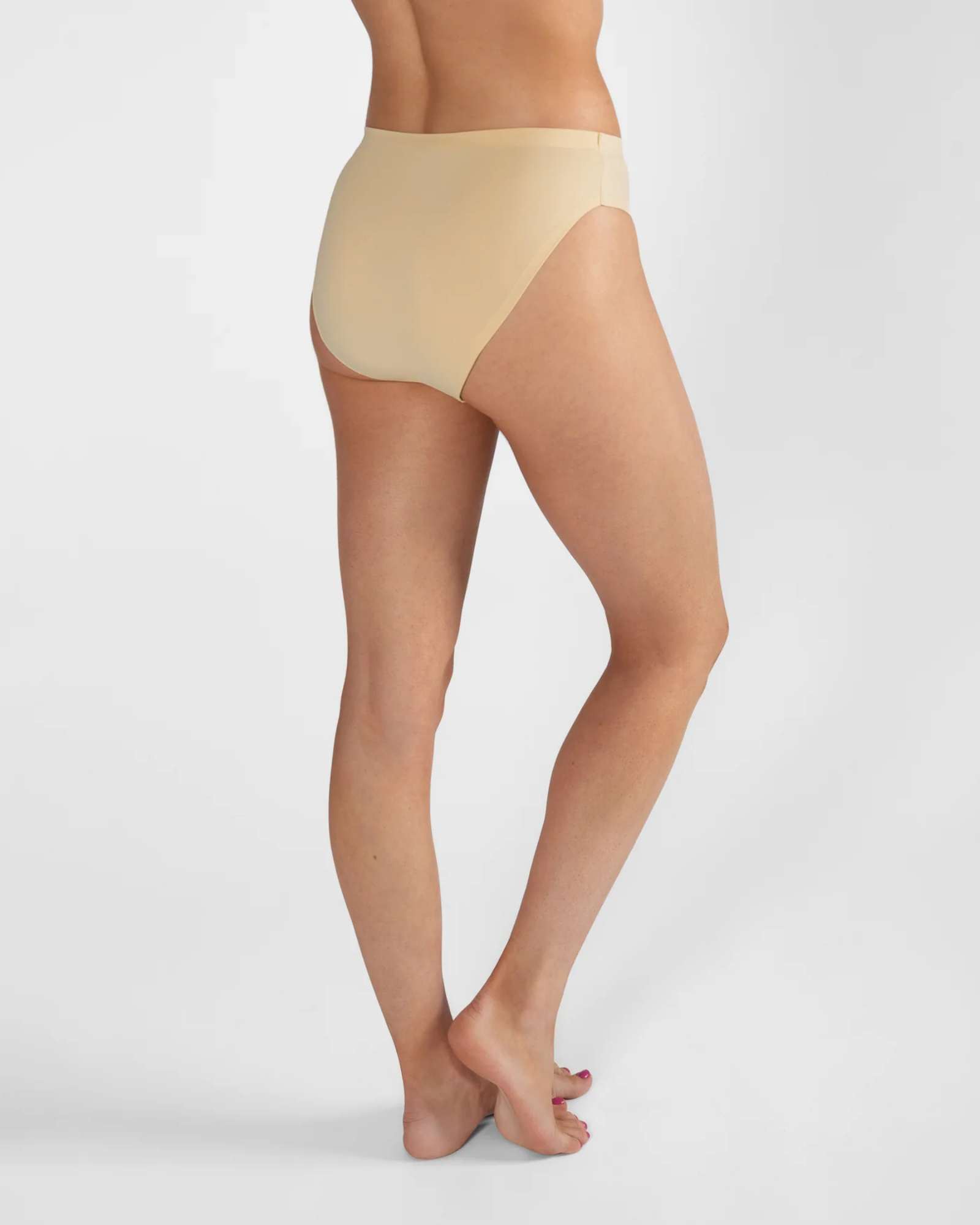 Rear three-quarter view of a model wearing high-waisted, seamless nude beige underwear against a light gray studio background.