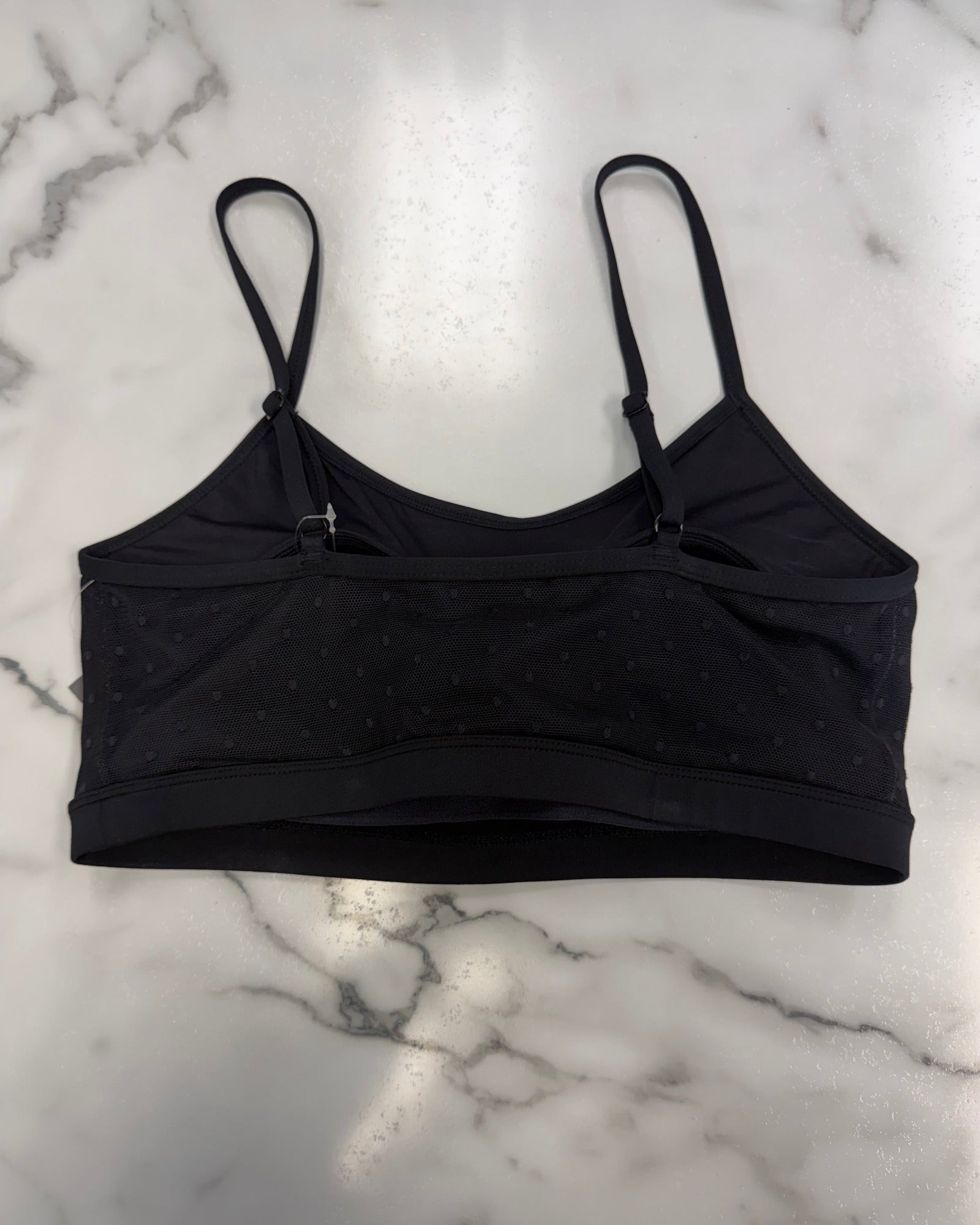 A black bra top with thin straps and a textured, dotted fabric pattern, laid flat on a white marble surface.