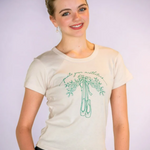 A smiling young woman wearing a cream-colored t-shirt with a green graphic of ballet slippers and the cursive text 'pointe your mistletoes'.