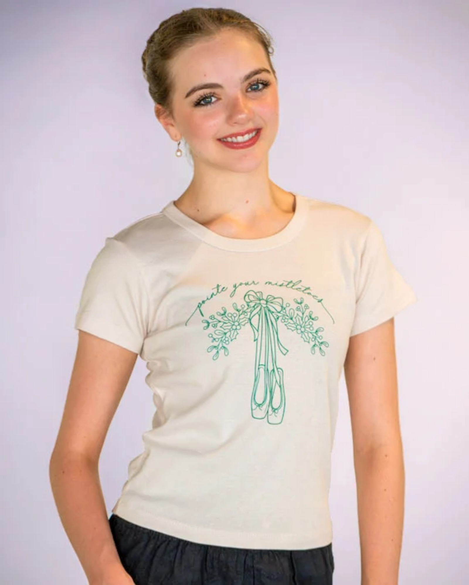 A smiling young woman wearing a cream-colored t-shirt with a green graphic of ballet slippers and the cursive text 'pointe your mistletoes'.