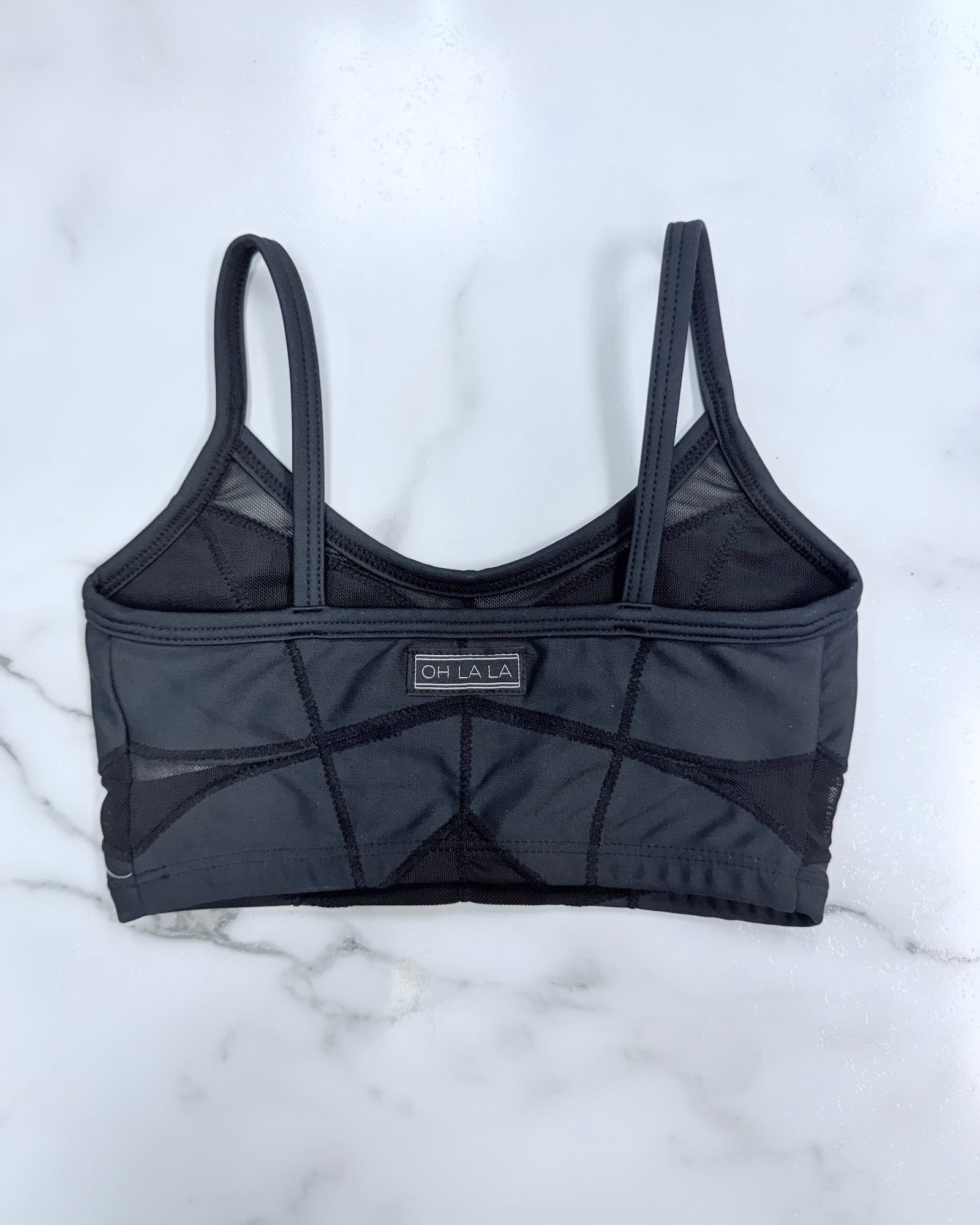 A dark gray mesh and structured sports bra with geometric paneling and a small 'CH LA LA' brand label on the front, laid flat on a white marble surface.