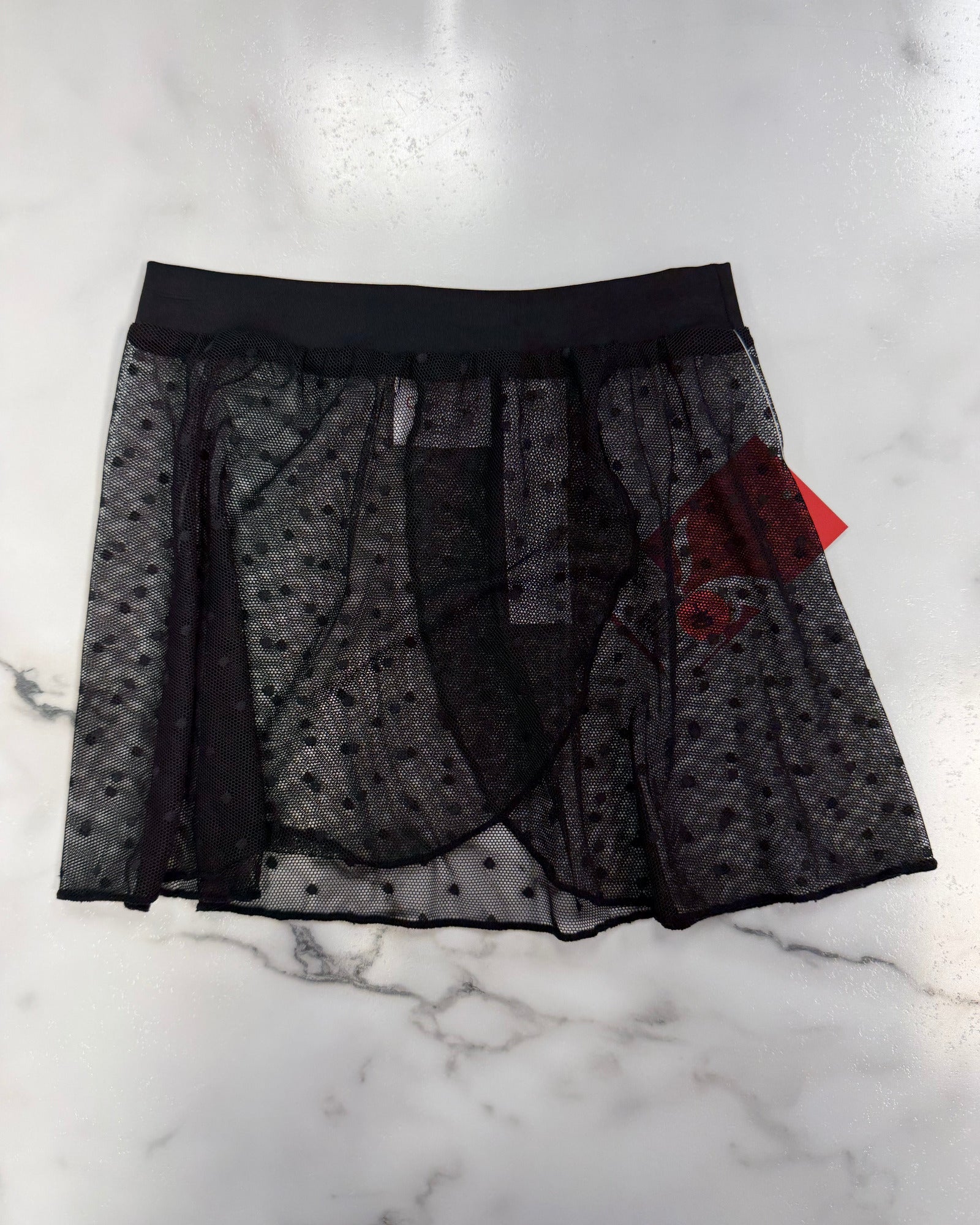 A sheer black mesh skirt with a subtle polka dot pattern, a solid black waistband, and a red brand tag, laid flat on a white marble surface.