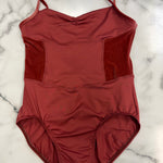 A rust-red leotard with thin shoulder straps and textured side panels, laid flat on a white marble surface.
