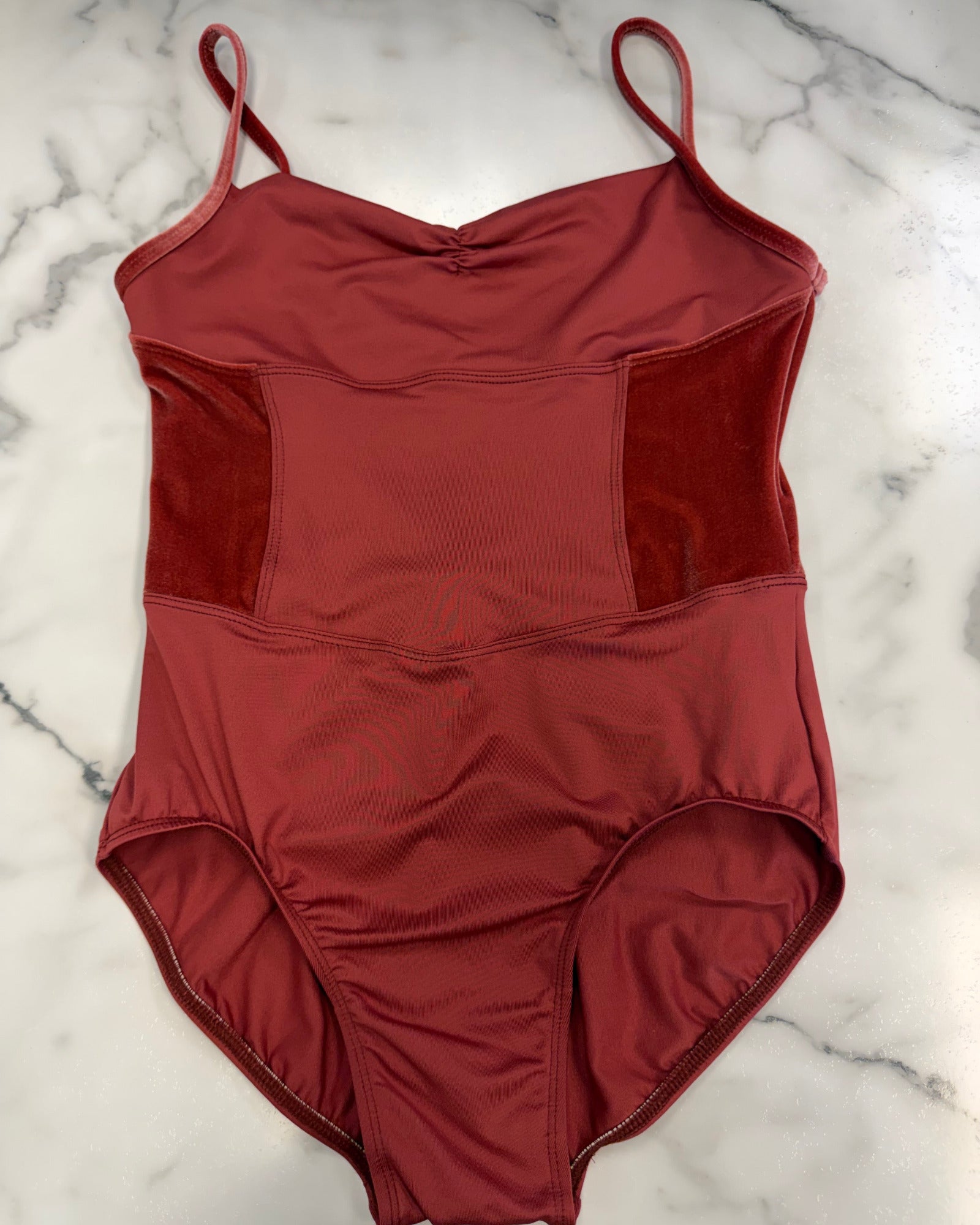 A rust-red leotard with thin shoulder straps and textured side panels, laid flat on a white marble surface.
