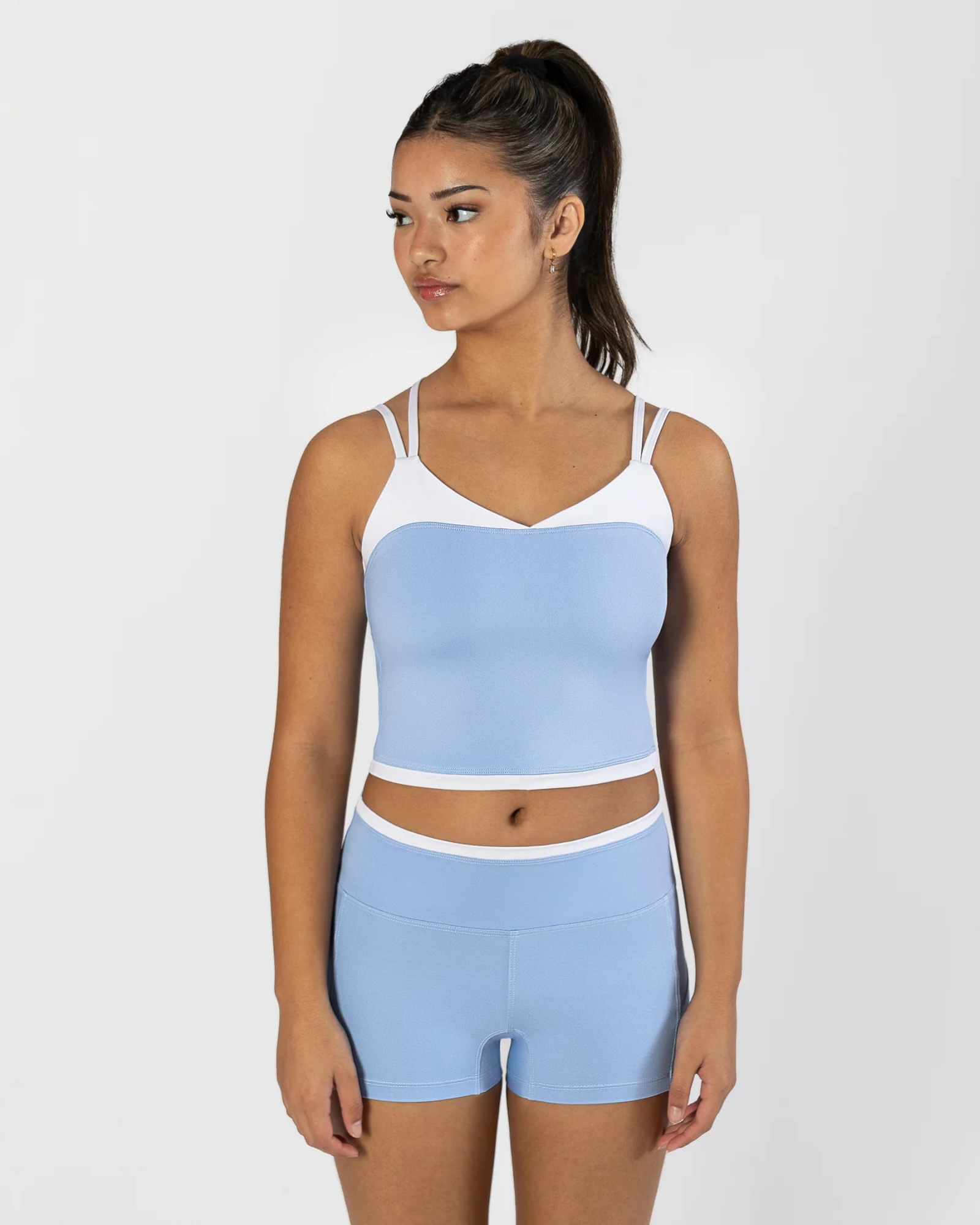 A woman modeling a light blue and white two-piece dancewear set, consisting of a cropped camisole top and matching high-waisted shorts, against a plain light gray background.
