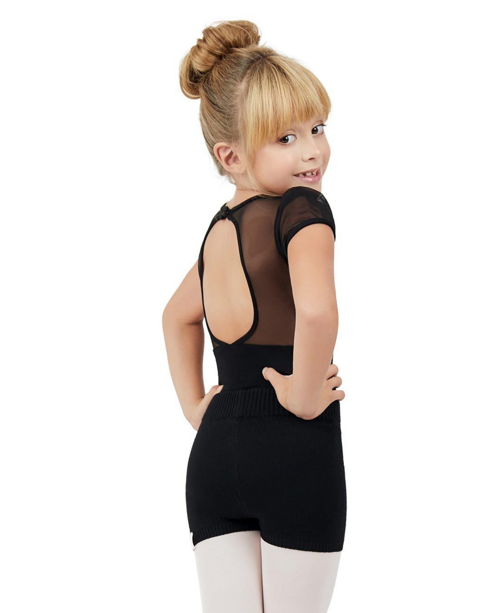 A young girl with blonde hair in a bun is shown from the back wearing a black short-sleeve leotard with sheer mesh back panels and white tights, set against a plain white background.