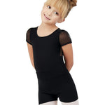 A young girl with blonde hair in a bun wearing a black dance leotard with sheer short sleeves and black dance shorts, paired with white tights, posing against a solid white background.