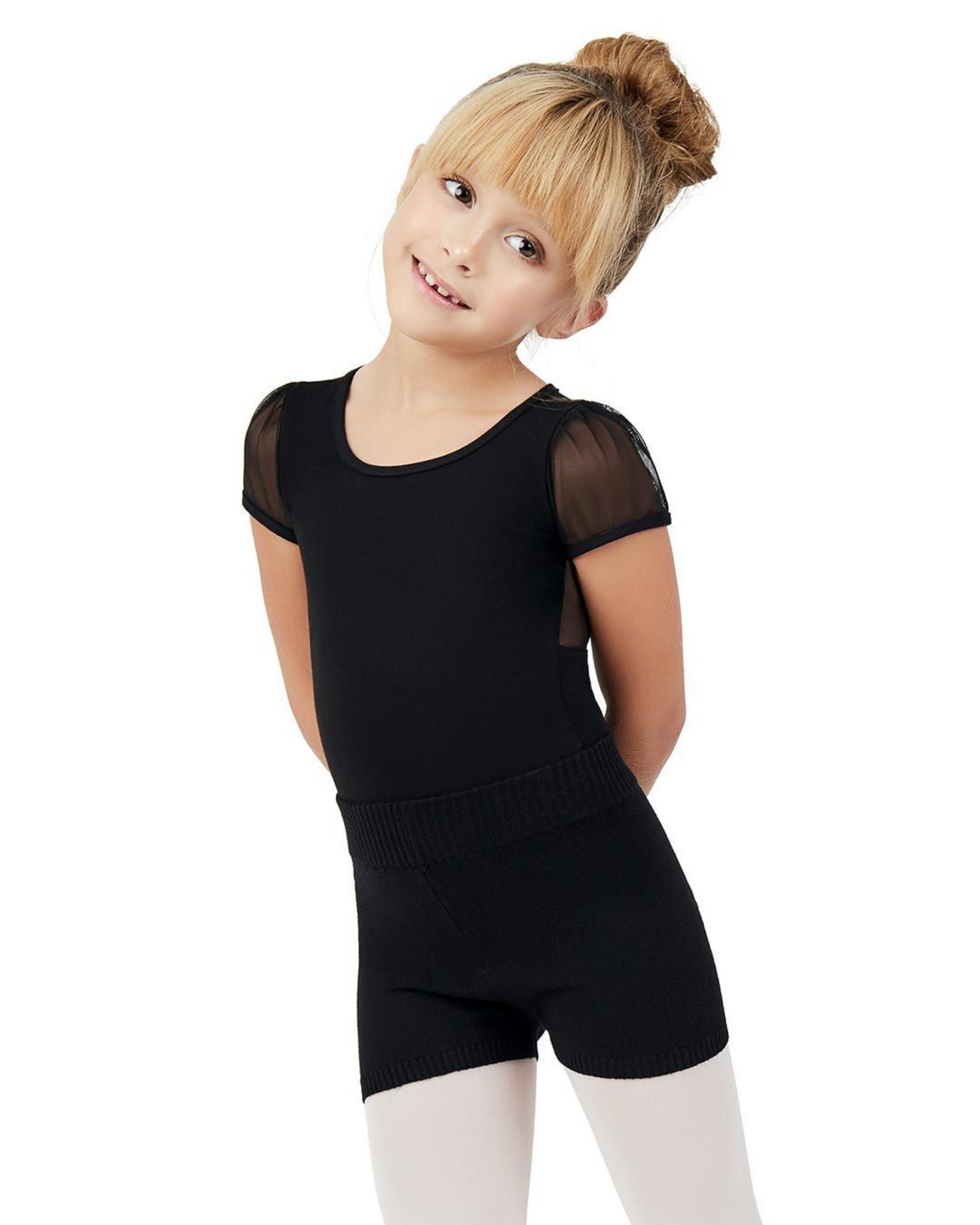 A young girl with blonde hair in a bun wearing a black dance leotard with sheer short sleeves and black dance shorts, paired with white tights, posing against a solid white background.