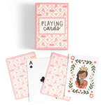 A boxed deck of playing cards with several cards fanned out below. The box and card backs have a pink patterned design with small illustrations, and one visible card shows a crowned queen illustration on a white background.