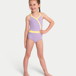 A young girl with braided hair stands barefoot against a light gray studio background, wearing a leotard in light purple with bright yellow trim.