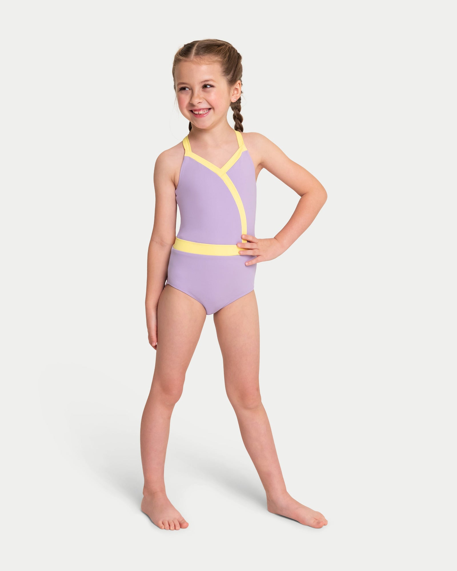 A young girl with braided hair stands barefoot against a light gray studio background, wearing a leotard in light purple with bright yellow trim.