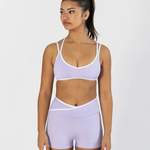 A woman modeling a light purple athletic set consisting of a dance bra and matching high-waisted shorts with white trim, set against a plain light gray background.