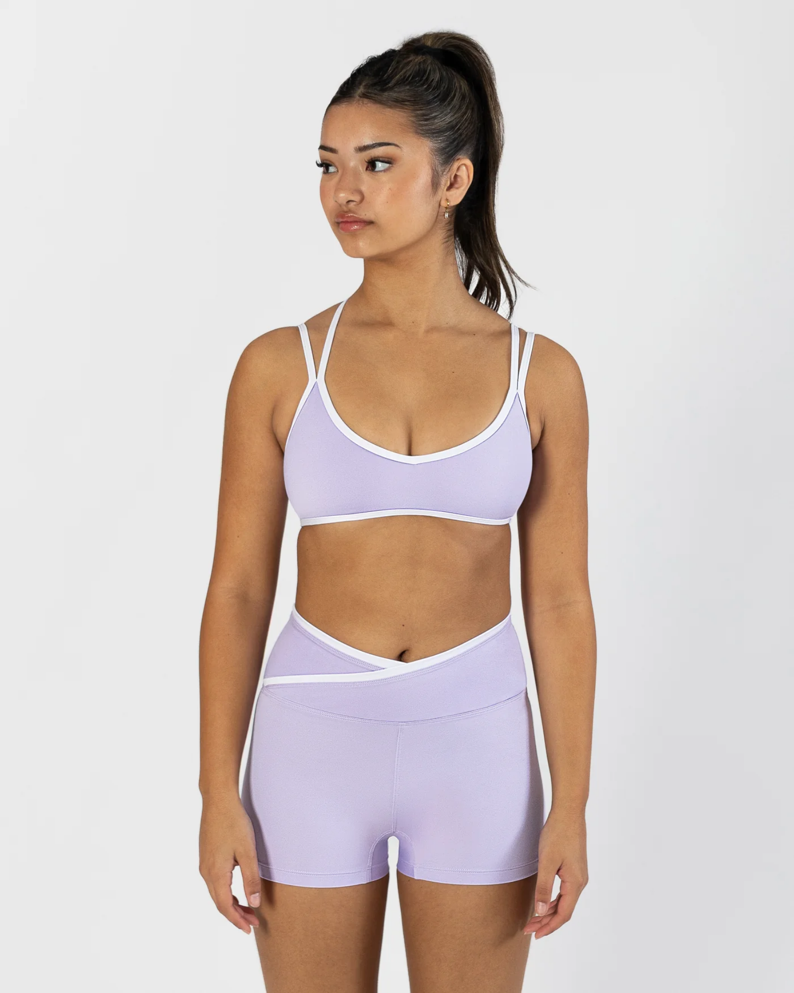 A woman modeling a light purple athletic set consisting of a dance bra and matching high-waisted shorts with white trim, set against a plain light gray background.