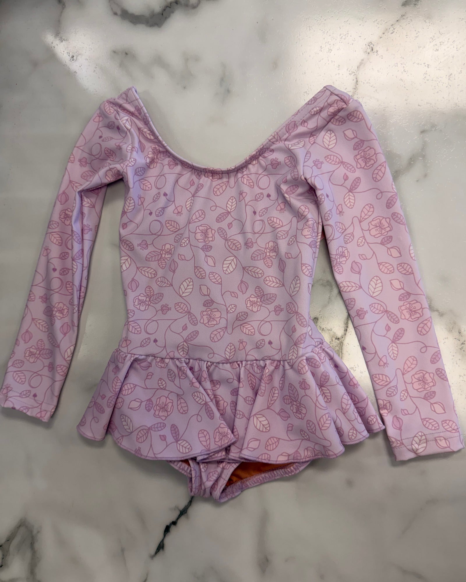 A long-sleeve baby/toddler leotard dress in light pink with a darker pink leaf pattern, laid flat on a white marble surface.
