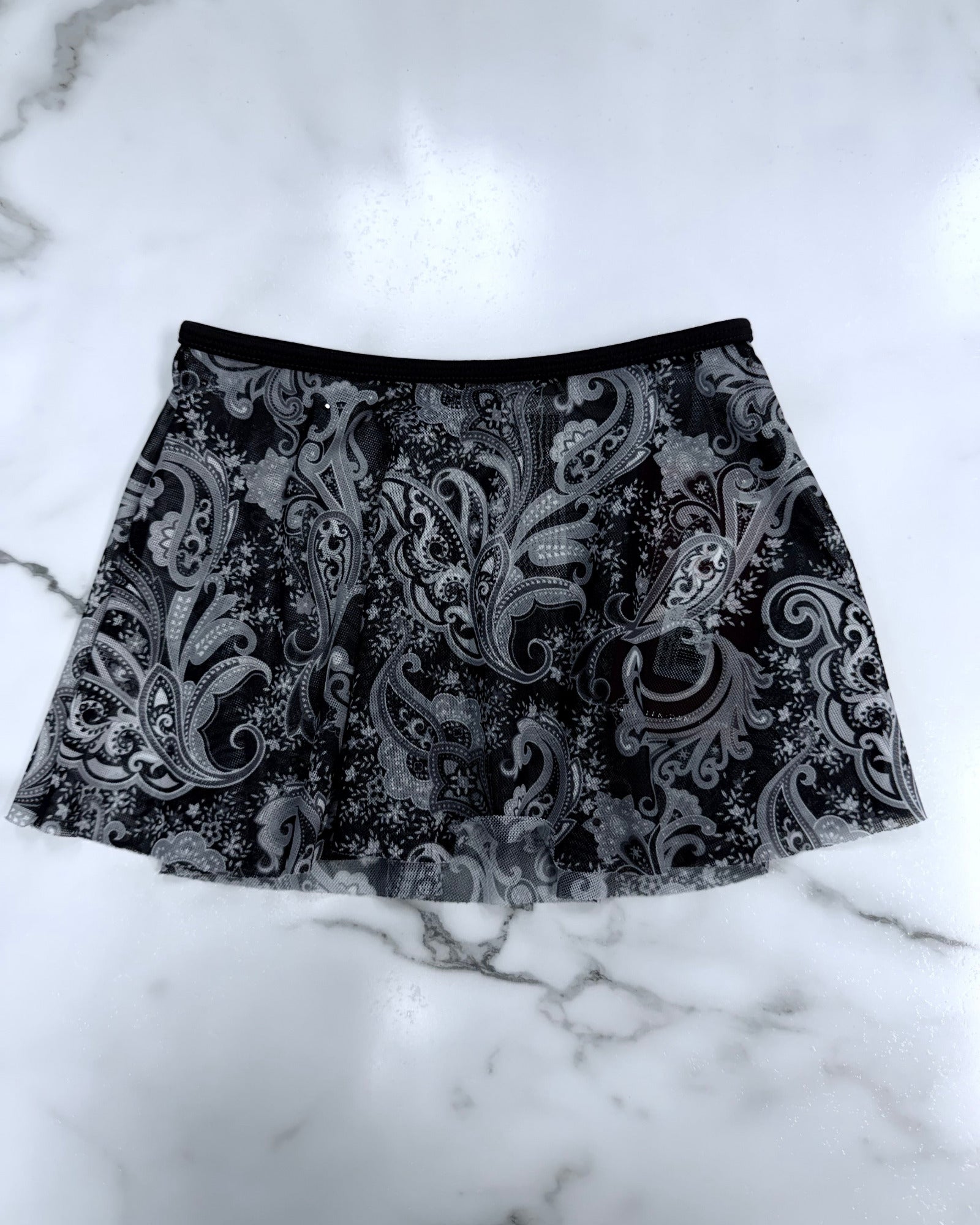 A black and gray paisley print dance skirt with a solid black waistband, laid flat on a white marble surface.