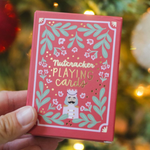 A hand holds a red box of Nutcracker playing cards decorated with green leaves, white flowers, and a nutcracker illustration, with a blurred Christmas tree in the background.