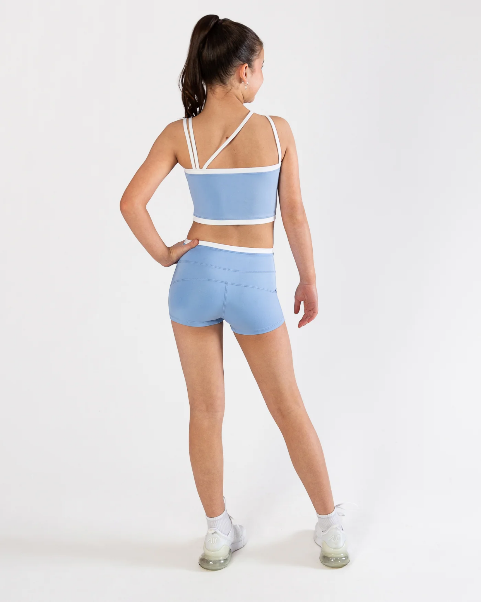 A girl modeling a light blue activewear set consisting of a dance bra and matching shorts, shown from a rear three-quarter view against a plain white studio background.