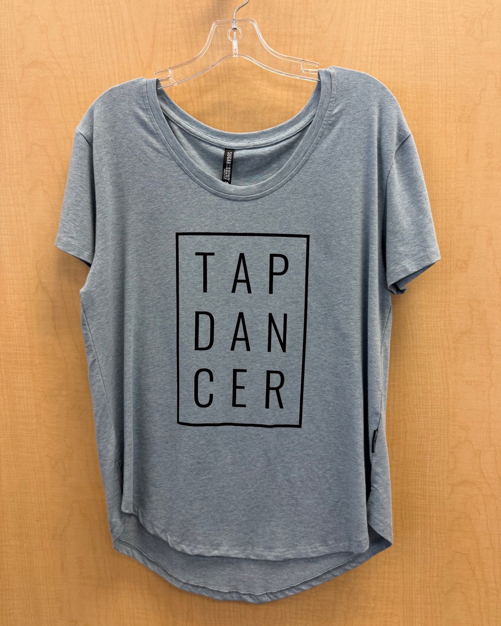 A heather blue short-sleeve t-shirt with a black graphic print reading 'TAP DANCER' inside a square, hanging on a clear plastic hanger against a light wood-paneled wall.