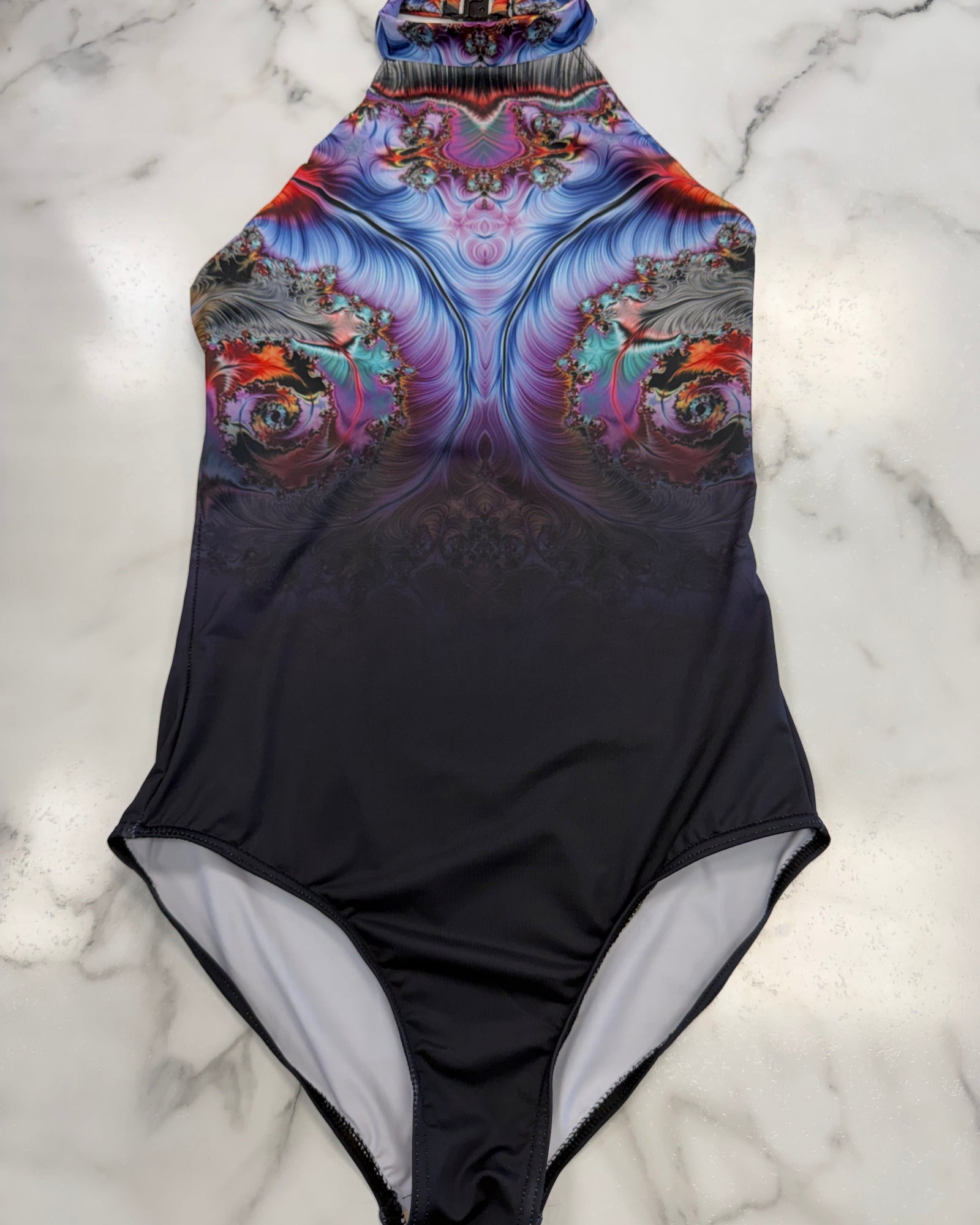 A one-piece halter leotard laid flat on a white marble surface. The leotard features a vibrant, symmetrical fractal floral pattern in shades of purple, blue, pink, and orange on the upper half, fading into a solid black lower half with a light gray interior lining.
