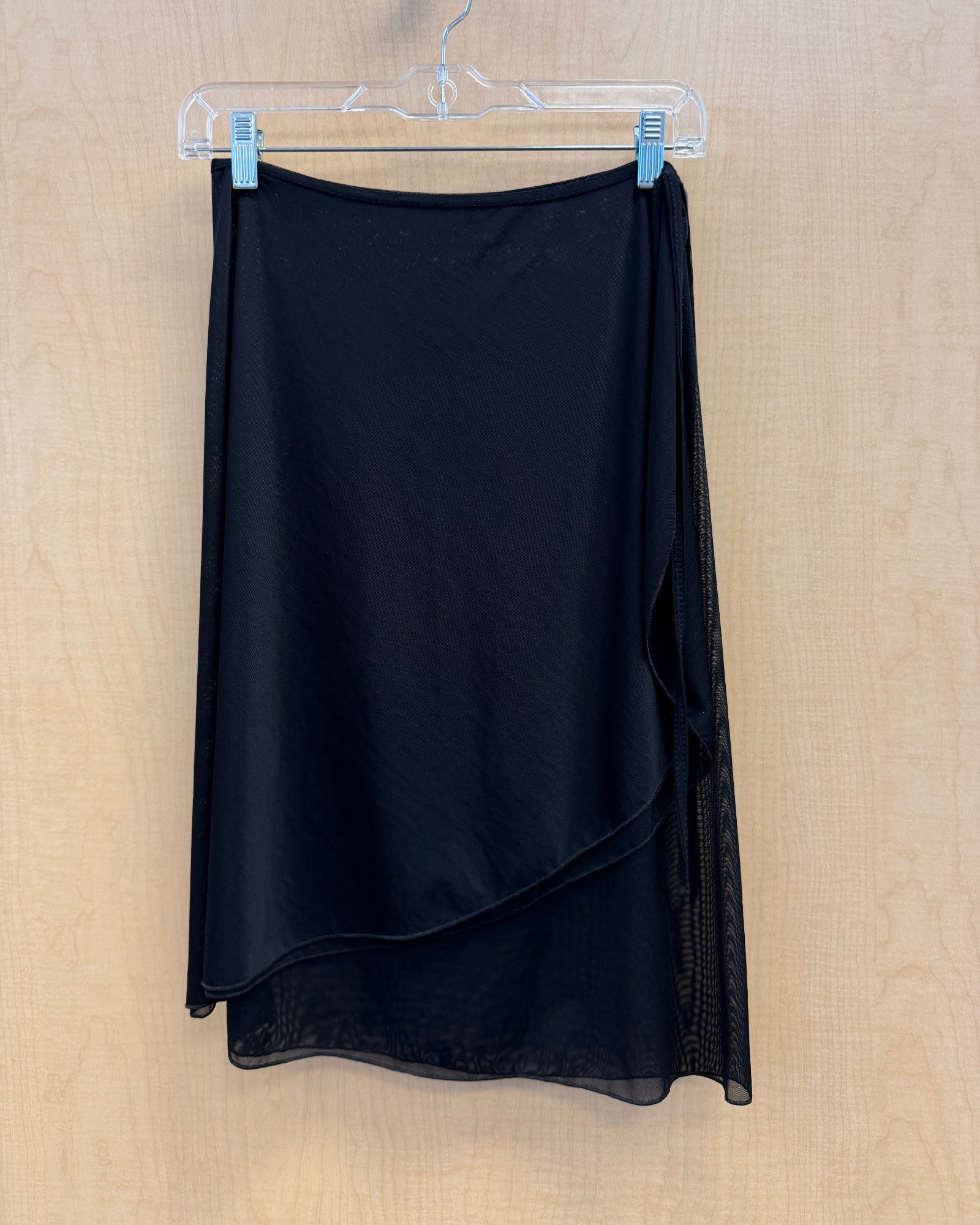 A black wrap-style midi skirt with a sheer mesh side panel, hanging on a clear plastic hanger against a light wood-paneled wall.