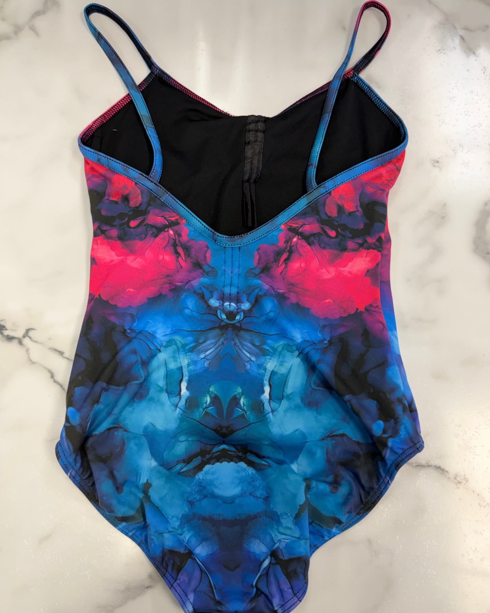 A leotard with a vibrant blue, pink, and black abstract watercolor pattern, laid flat on a white marble surface.