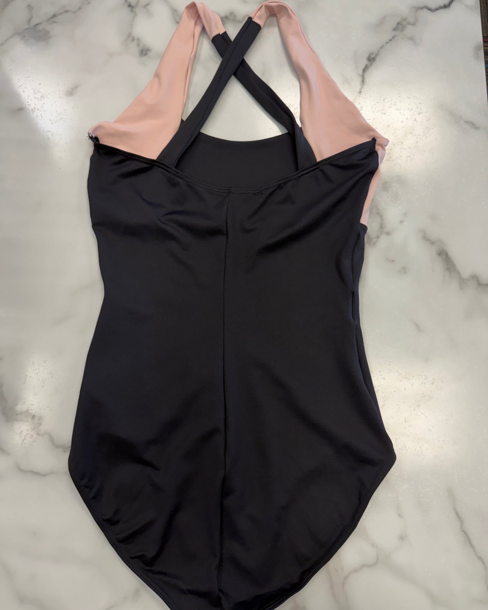 A black one-piece leotard with light pink shoulder straps, laid flat on a white marble surface.