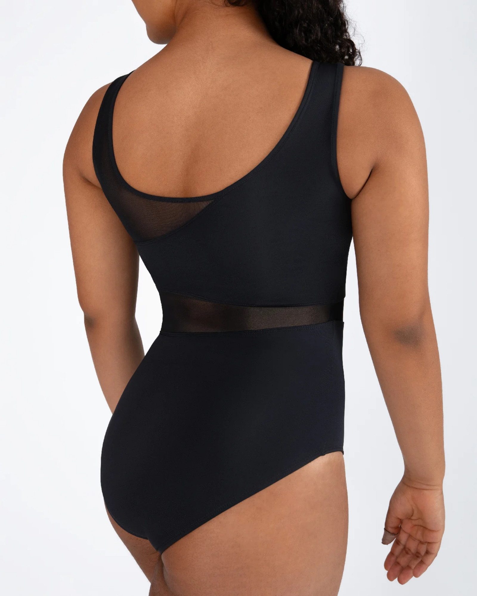 A woman shown from the back wearing a black one-piece leotard with sheer mesh panels at the back and around the waist, photographed against a light gray studio background.