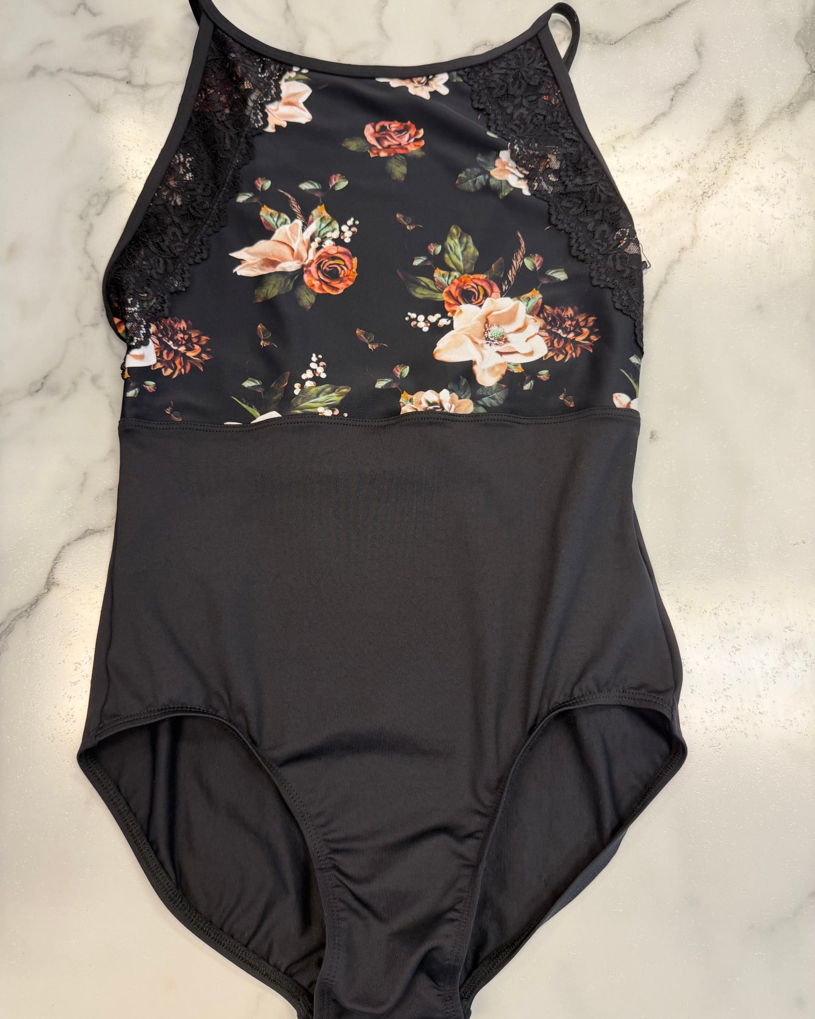 A black one-piece leotard with a floral print bodice and lace trim, laid flat on a white marble surface.