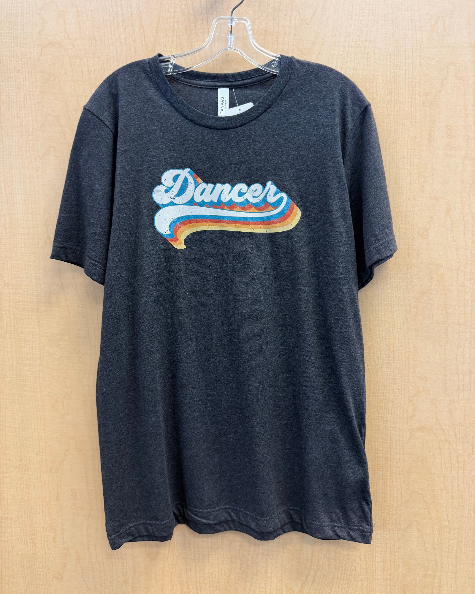 A dark charcoal gray heather t-shirt hanging on a clear plastic hanger against a light wood-paneled wall. The shirt features a retro-style graphic on the chest with the word 'Dancer' in white script over a wavy blue, orange, and yellow stripe design.