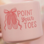 A close-up of a light pink ceramic mug held by a hand, featuring a pink ballet shoe illustration and the text 'POINT YOUR TOES' in pink lettering.