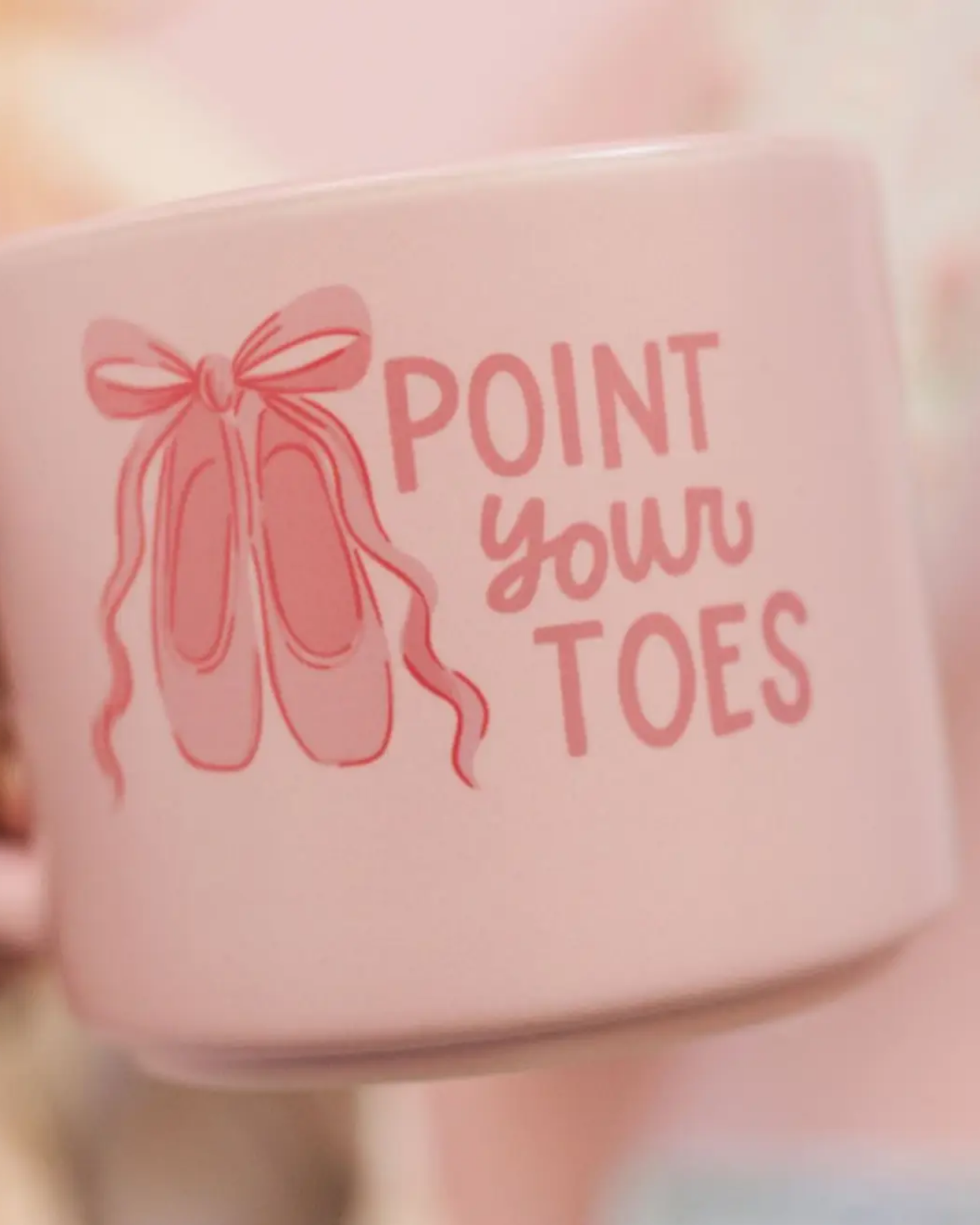 A close-up of a light pink ceramic mug held by a hand, featuring a pink ballet shoe illustration and the text 'POINT YOUR TOES' in pink lettering.